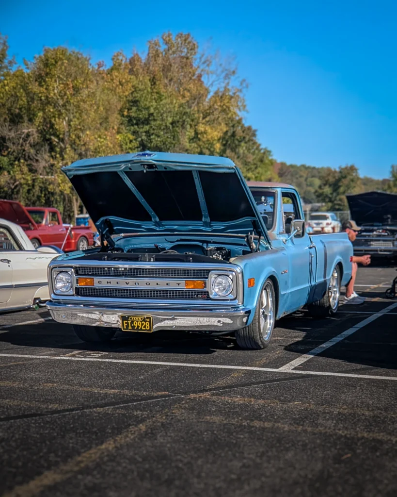 A light blue Chevrolet C10 pickup sits with its hood raised in a parking lot at an outdoor car show.