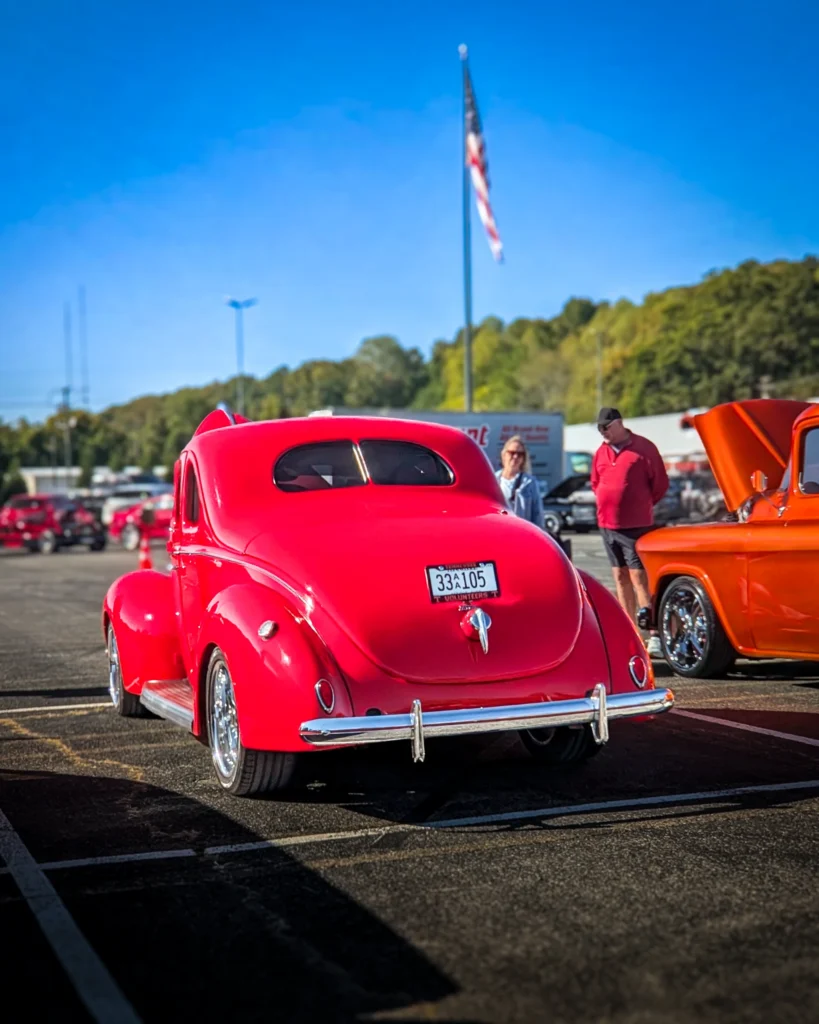 Red Ford Deluxe Coupe parked at car event with an orange truck partially visible on the right and people in background