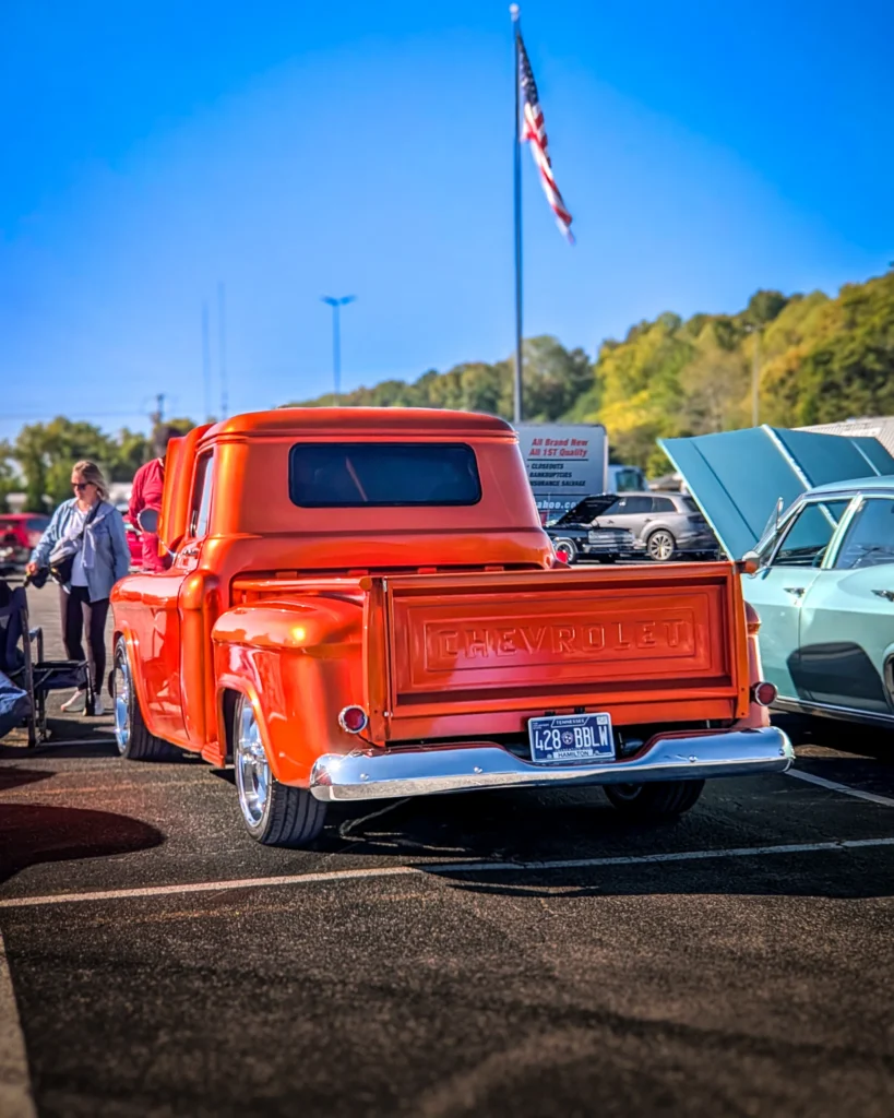 Rear view of orange Chevrolet 3100 stepside parked outdoors with open-hood light blue car next to it under clear sky.