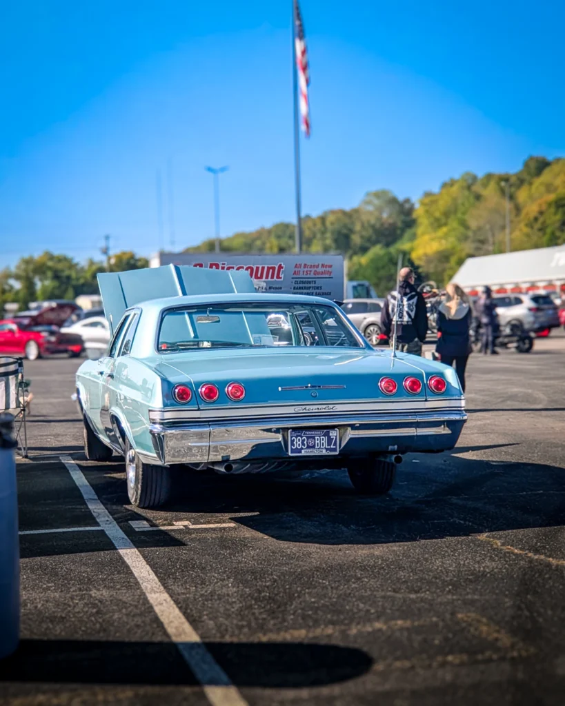 A light blue Chevrolet Impala sits with its hood raised in a car show parking lot, viewed from the rear.