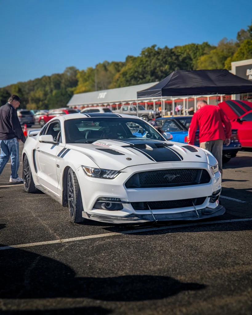 White Ford Mustang GT with black racing stripes parked at an outdoor car event with attendees nearby under a clear blue sky.