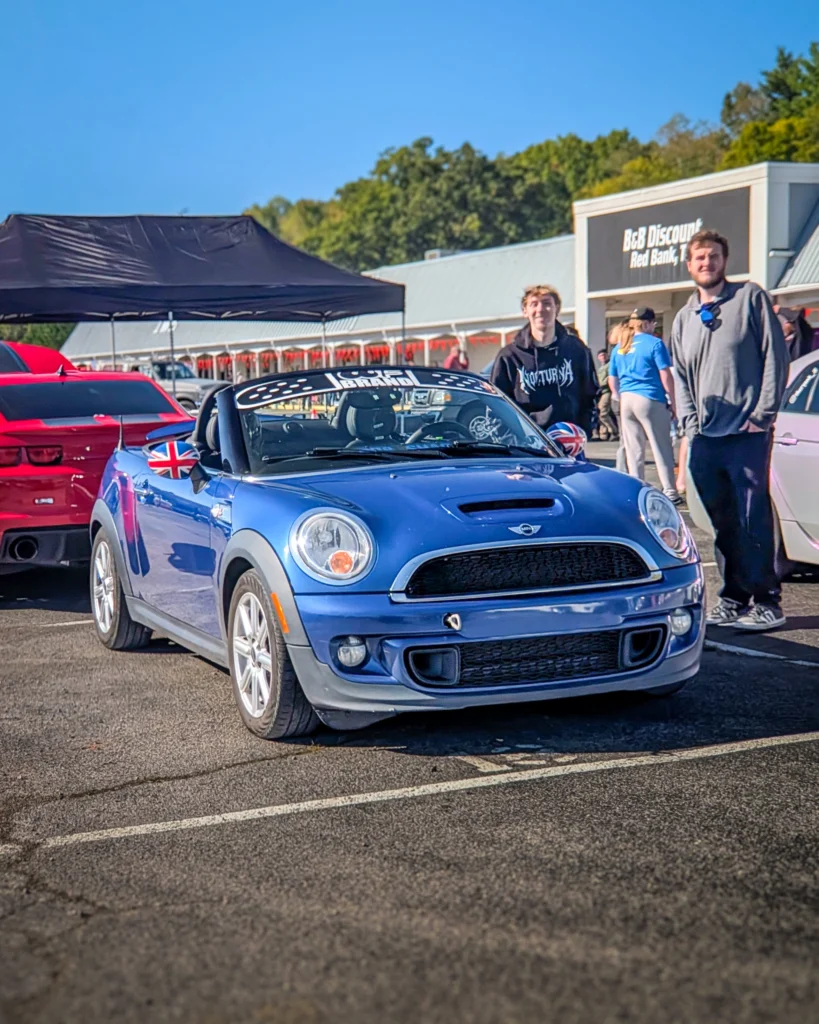 Blue Mini Cooper convertible with Union Jack mirror covers parked outdoors at a car event with two people standing nearby.