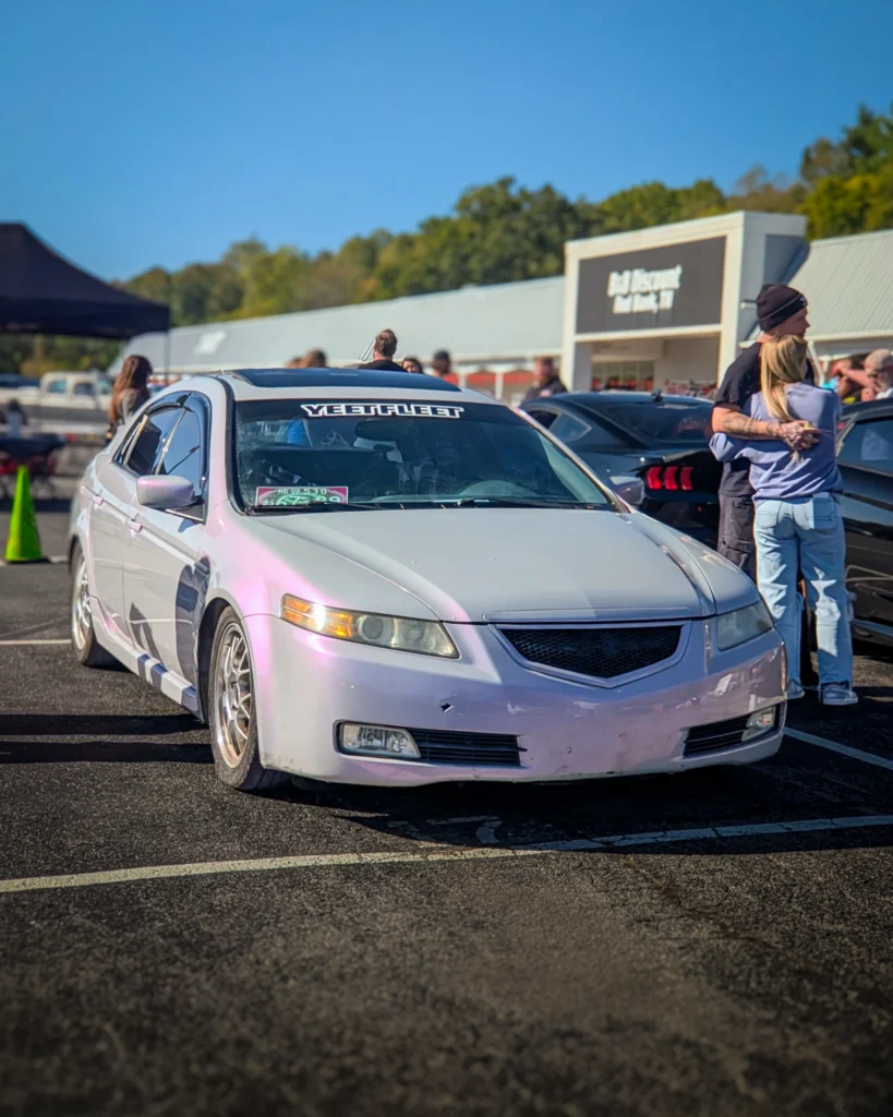 A silver Acura TL with pink reflections is parked beside attendees and other cars at an outdoor car meet.