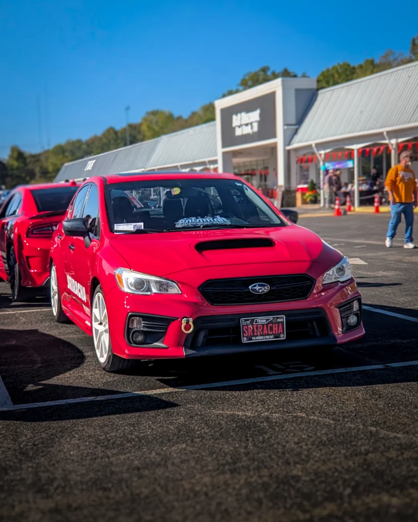 Red Subaru sedan with hood scoop parked in a lot at a car event with another red car behind it and people near a building