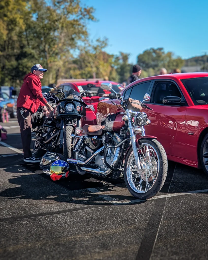 A burgundy motorcycle and a black motorcycle are parked beside red cars, with an attendee and helmets nearby.