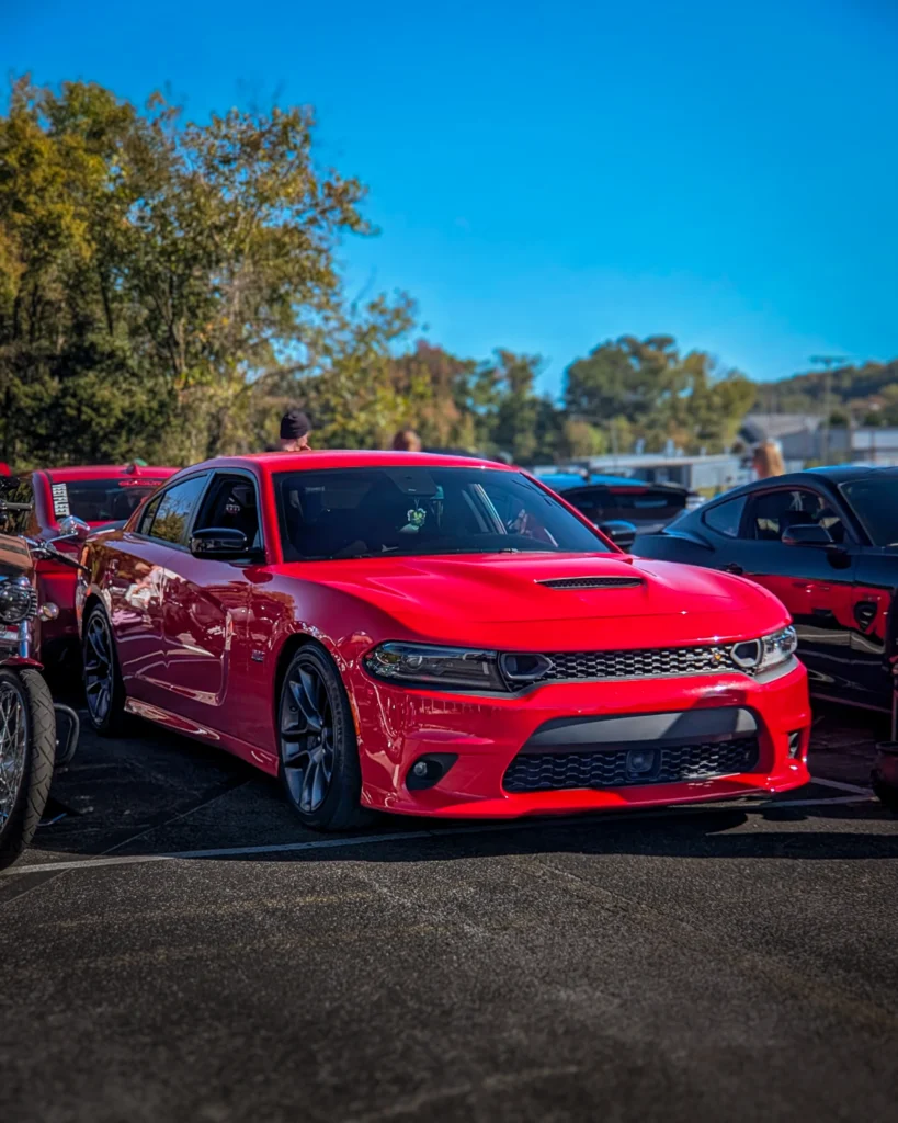 Red Dodge Charger parked outdoors at car event with trees and people in background under clear blue sky