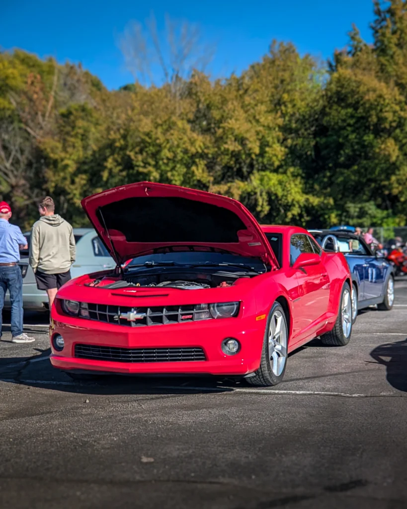 Red Chevrolet Camaro with hood open parked at an outdoor car event with people and other vehicles in the background.