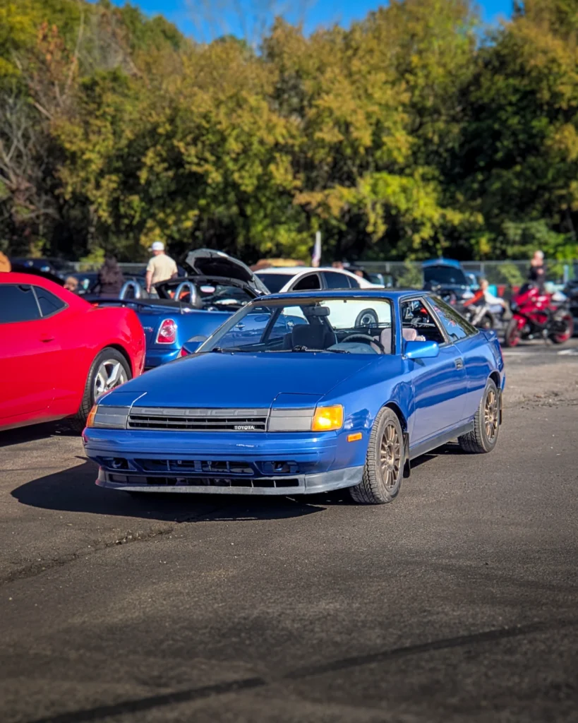 Blue Toyota Celica parked outdoors at a car event with other vehicles and people in the background