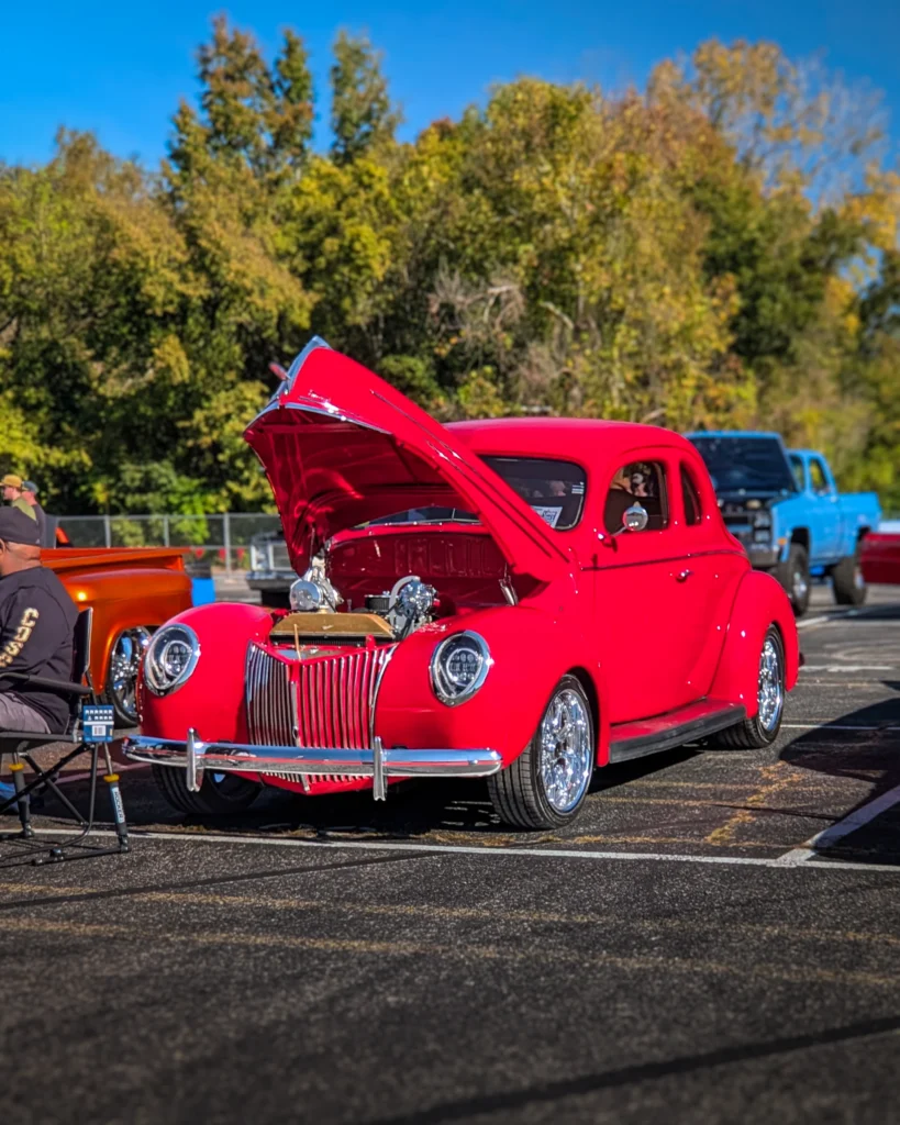 Ford Deluxe Coupe with open hood parked outdoors with trees and other vehicles in the background