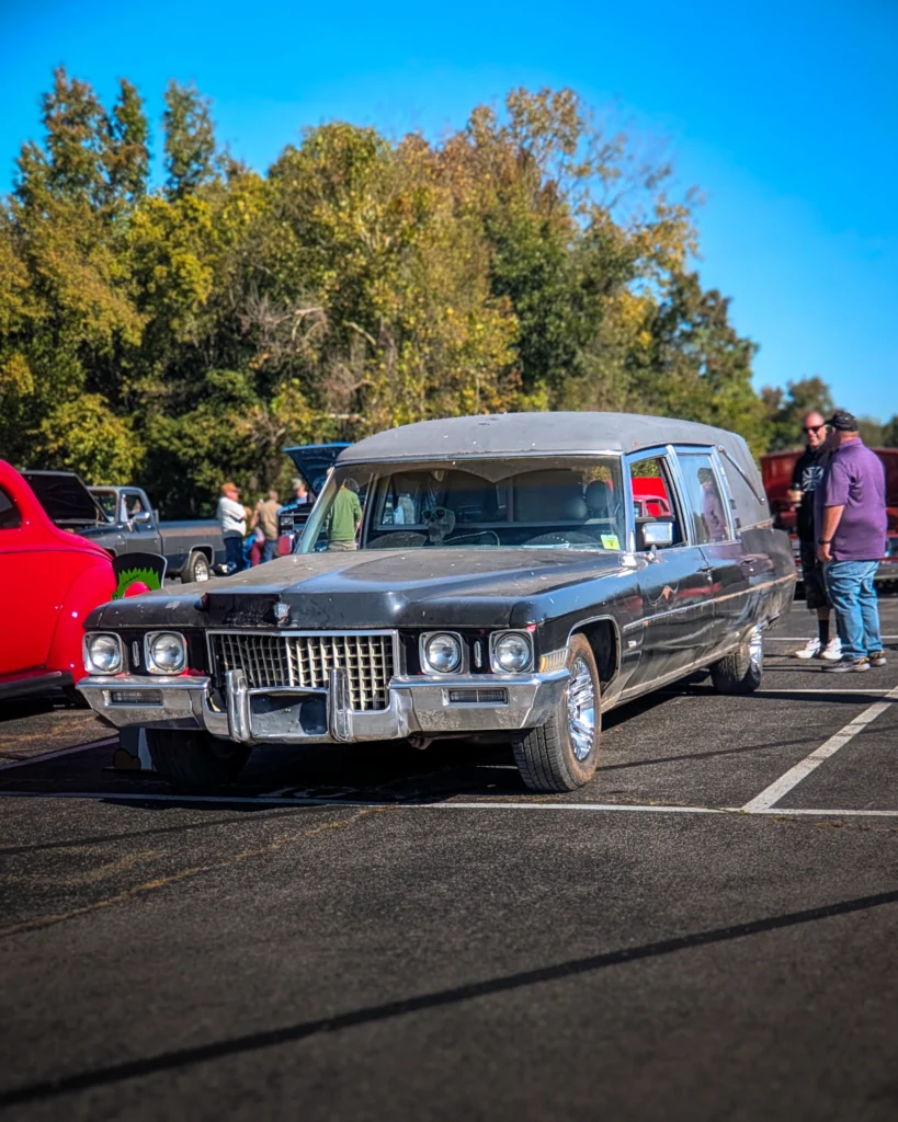 Black vintage Cadillac hearse parked at a car event with people and other vehicles in the background under clear blue sky.