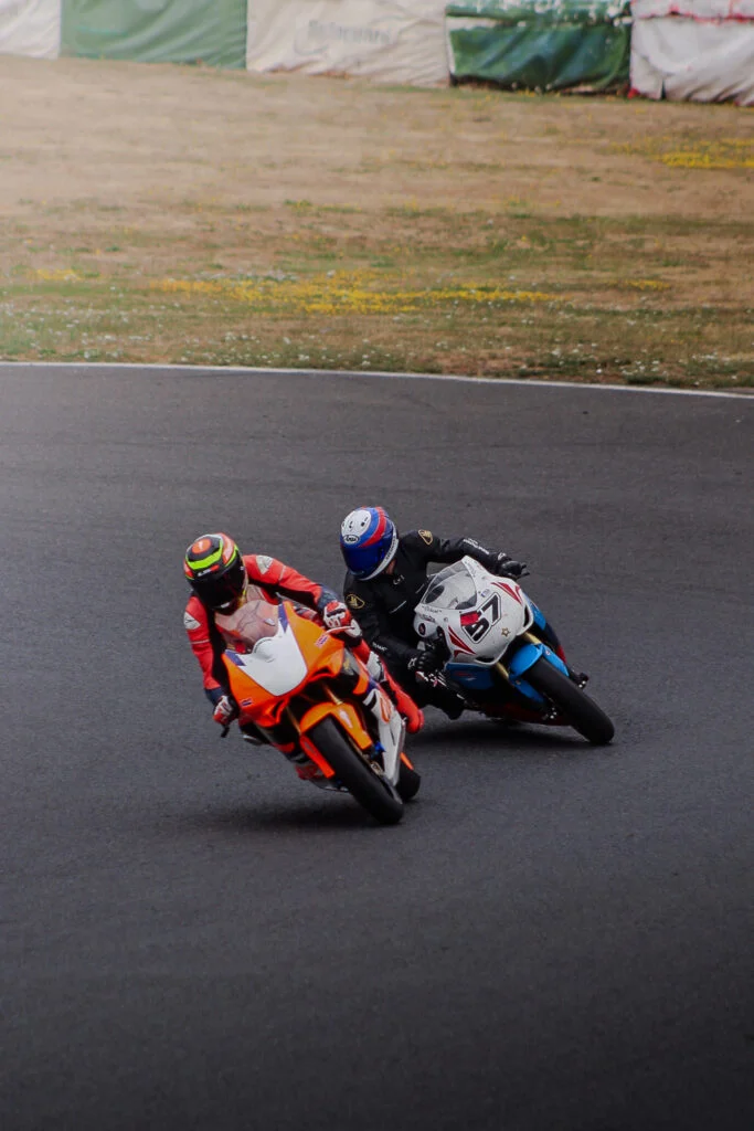 Two motorcyclists competing on a racetrack, leaning into a curve, one on an orange bike and the other on a white and blue bik