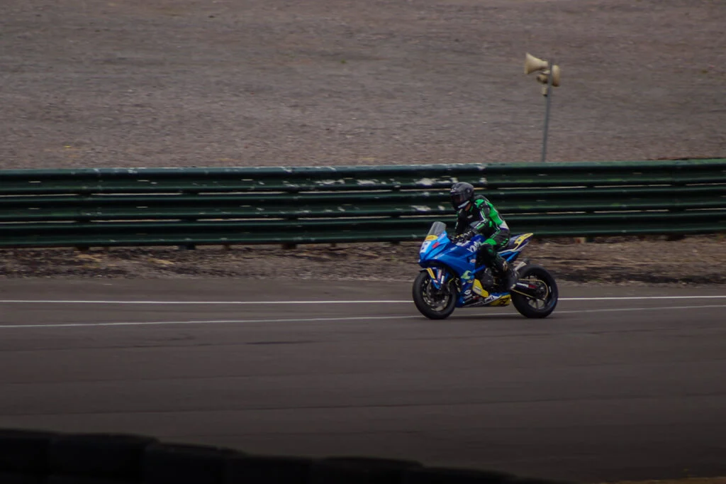 Blue racing motorcycle with rider in black and green suit on a paved track beside a green metal barrier