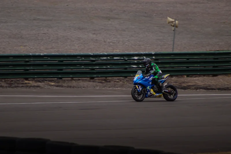 Blue racing motorcycle with rider in black and green suit on a paved track beside a green metal barrier