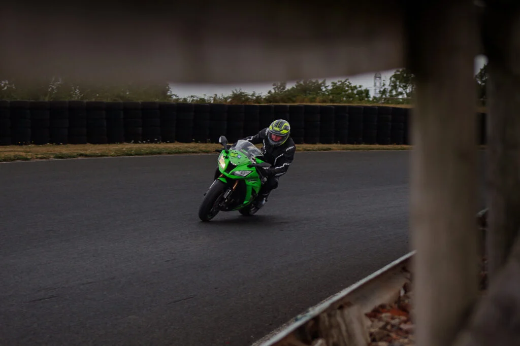 Motorcycle rider in black gear on green Kawasaki sportbike leaning into a curve on a racetrack with tire barriers in backgrou