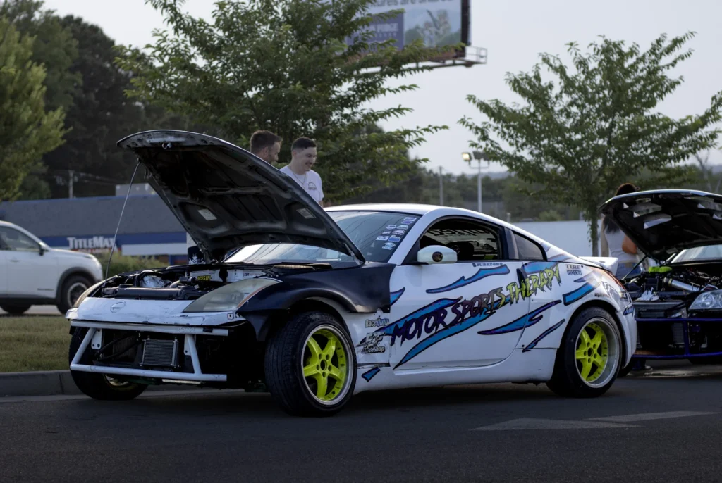 White Nissan 350Z with hood open and neon yellow wheels at a car meet with people and trees in the background