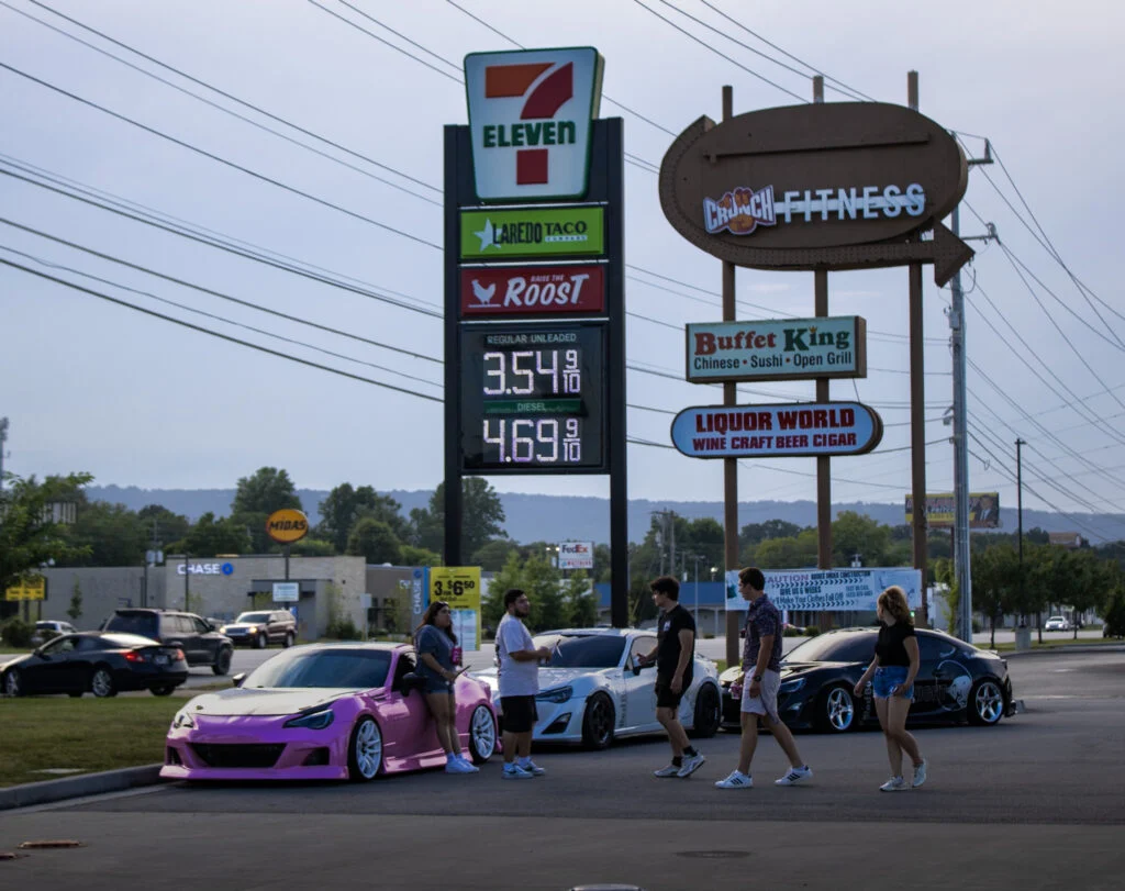 Group of people near three parked sports cars in a parking lot with commercial signage in the background
