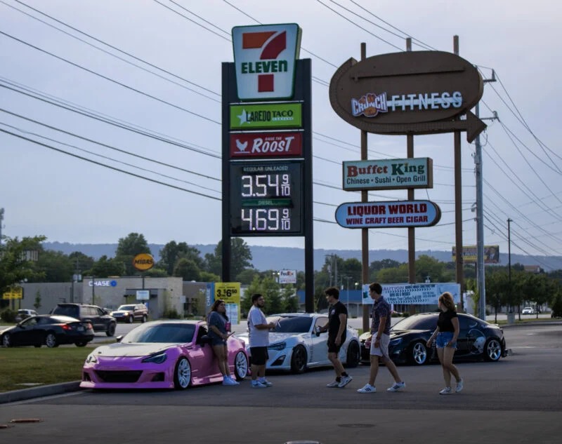 Group of people near three parked sports cars in a parking lot with commercial signage in the background