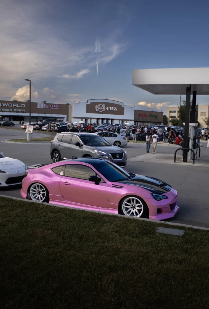 Pink Subaru BRZ with white wheels parked near a gas station canopy in front of a crowded shopping plaza parking lot