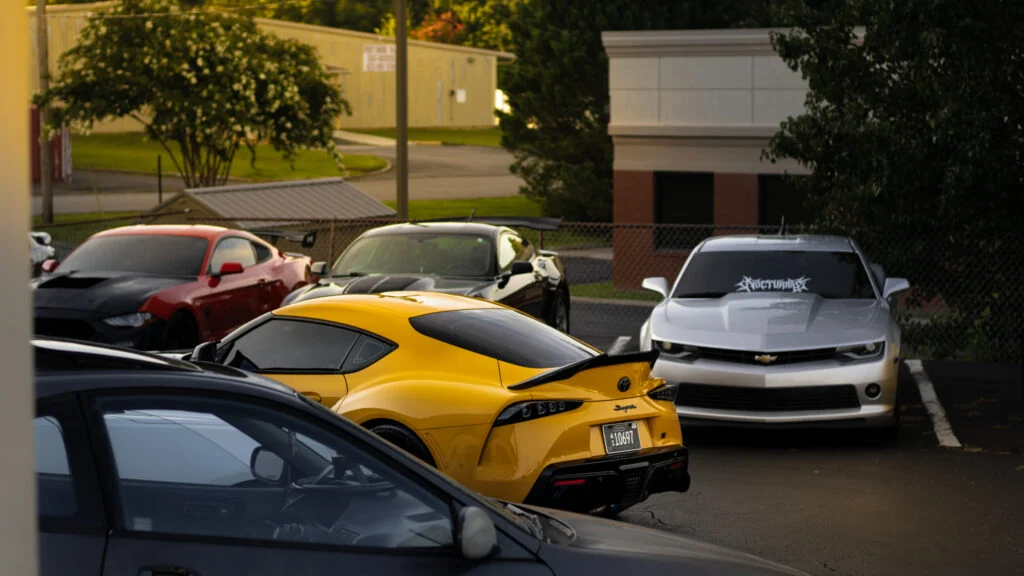 Yellow Toyota Supra, red Ford Mustang, black car, and white Chevrolet Camaro parked in a lot near buildings and trees