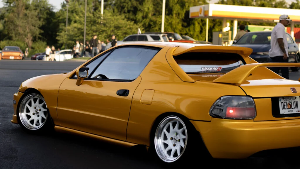 Yellow Honda Civic Del Sol with white wheels parked near a gas station with people in the background