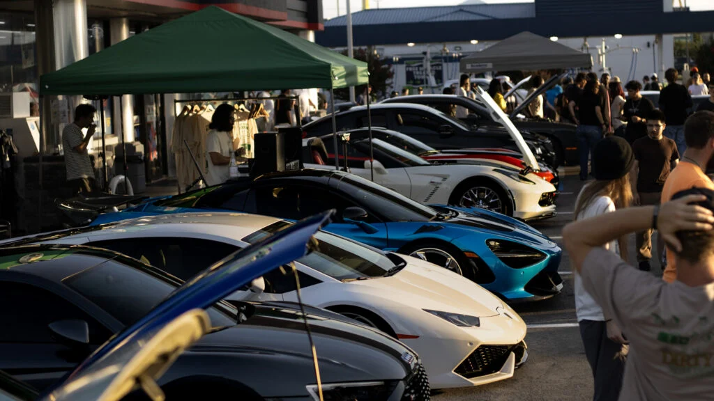 Row of sports cars including white Lamborghini and blue McLaren at an outdoor car event with people and tents in background