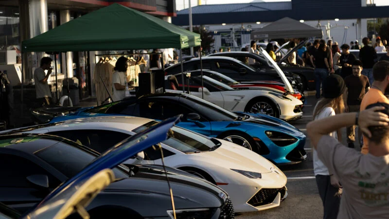 Row of sports cars including white Lamborghini and blue McLaren at an outdoor car event with people and tents in background