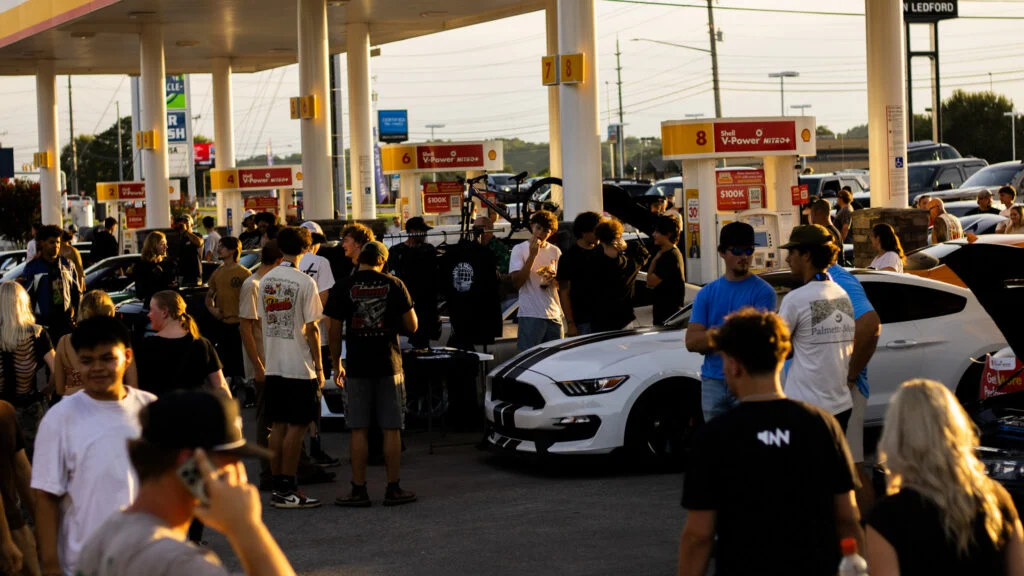 Crowd of people gathered at a Shell gas station around parked cars including a white Ford Mustang with black stripes.