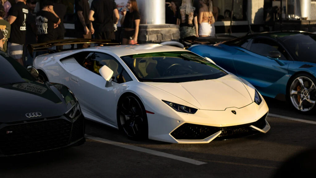 White Lamborghini Huracán with black rear wing and black wheels parked next to a black Audi and a blue sports car with people