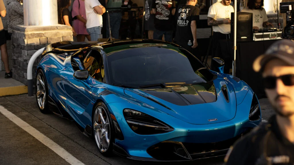 Blue McLaren 765LT parked on a street with people and a DJ setup in the background