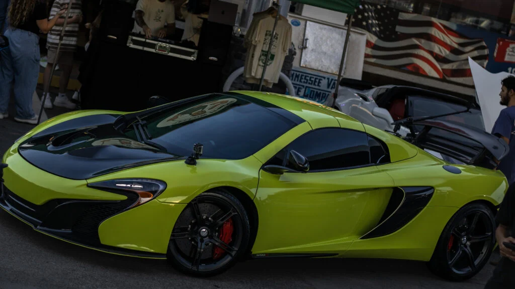 Green McLaren 650S parked at a car event with people and an American flag in the background