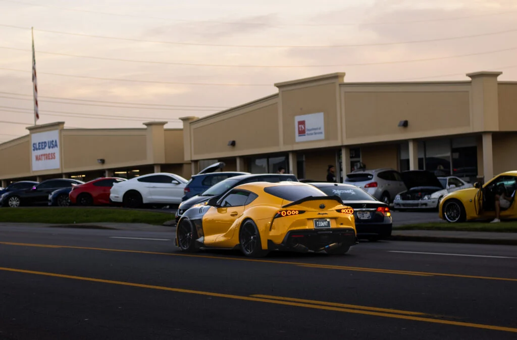 Yellow Toyota Supra coupe driving on a street near other parked cars and commercial buildings during sunset