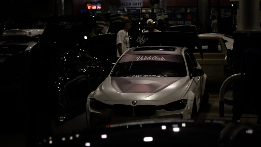 White BMW F30 with 'Valid Club.' sticker on windshield parked indoors among other cars and people