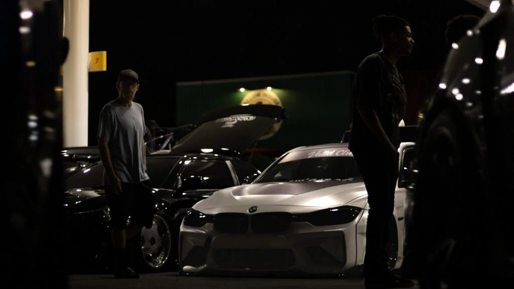 Two men standing near a white BMW F30 with hood open at night under artificial lighting at a car meet