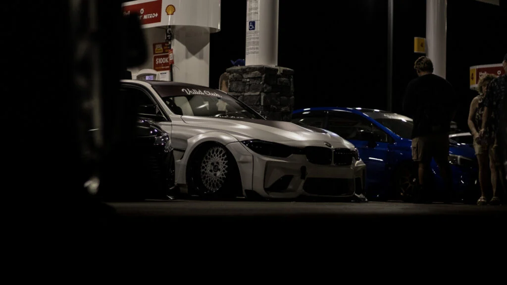 White BMW F30 with aftermarket wheels parked at a gas station at night with other cars and several people nearby.