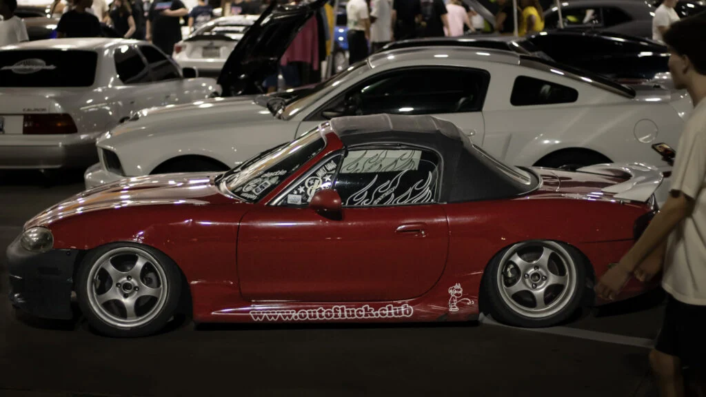 Red Mazda MX-5 Miata with black soft top and decals on windows and sides parked alongside white vehicles at a nighttime event