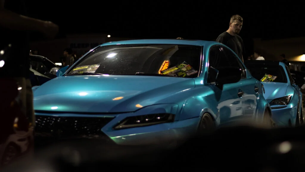 Two teal sedans parked outdoors at night with a man standing near the second car, reflected light on cars' surfaces