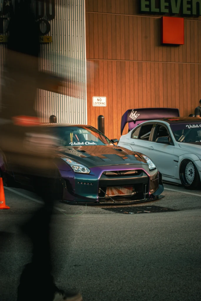 Purple Nissan GT-R with front windshield decal reading 'Valid Club' next to a white car with a large purple spoiler in a park