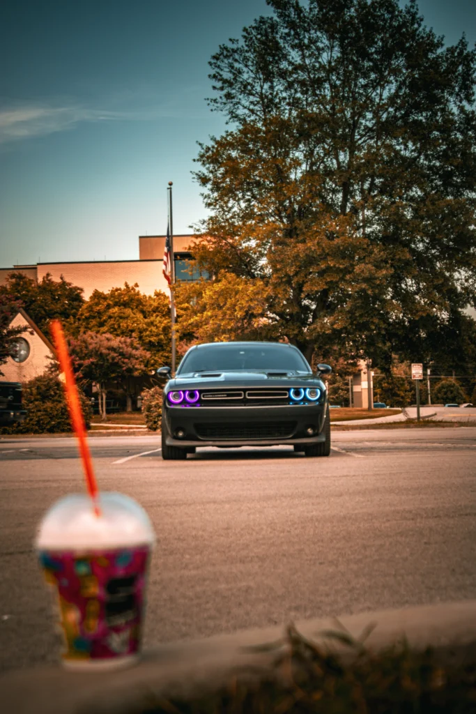 Black Dodge Challenger with purple and blue halo headlights parked outdoors near trees and buildings during daytime