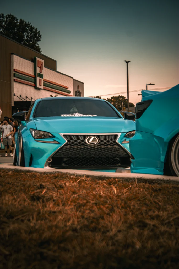 Light blue Lexus RC 350 parked near a 7-Eleven with people and streetlights in the background at dusk.