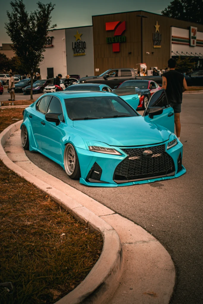 Bright blue Lexus IS F-Sport parked near a curb with a person standing beside the open driver door at a car meet parking lo