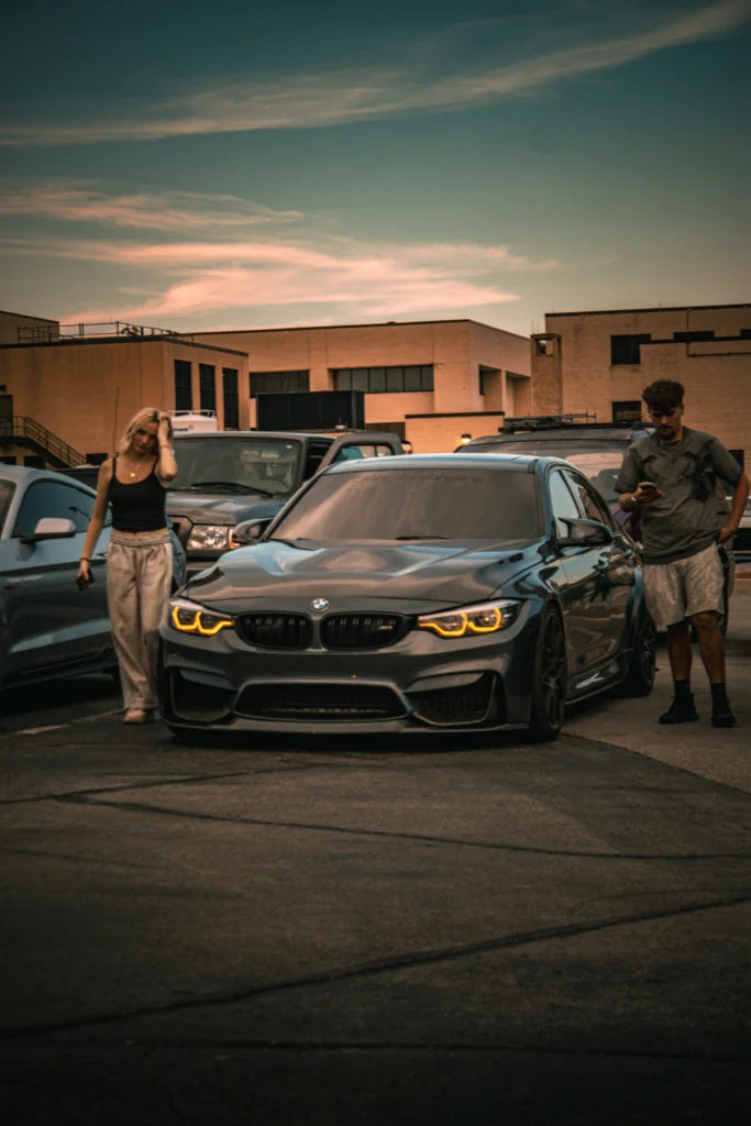 Black BMW F80 M3 with headlights on among other parked vehicles and people standing near it at dusk by brick buildings