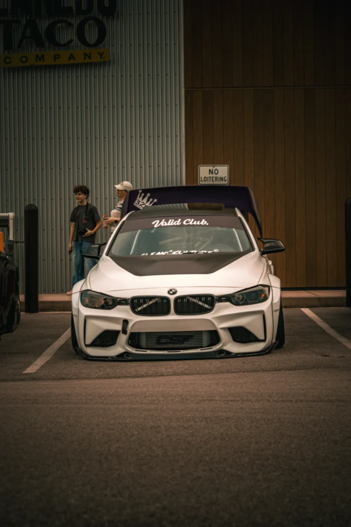White BMW 3 Series with black hood and large rear wing parked in front of a building with corrugated metal and wood walls