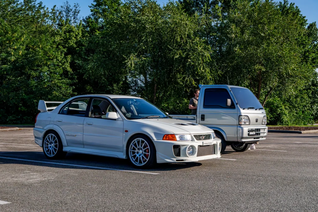 White Mitsubishi Evolution VI sedan parked next to a silver Honda Acty mini truck in a sunlit parking lot with trees in backg