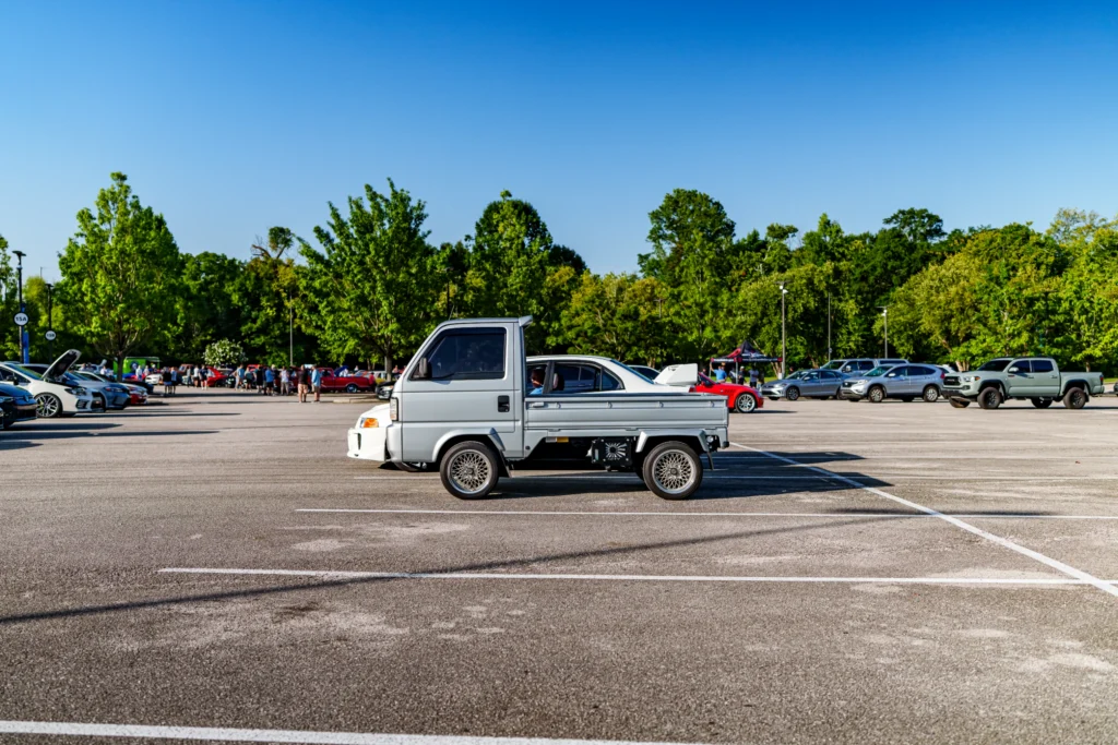 Silver Honda Acty kei truck parked in a lot with a car meet crowd and trees in the background under blue sky