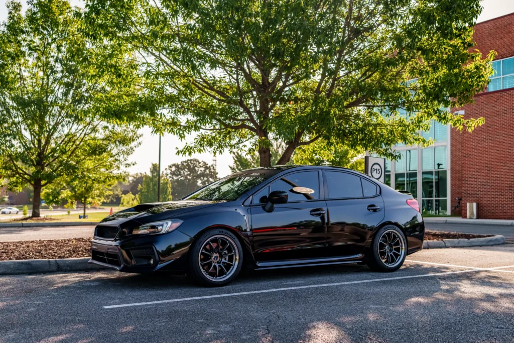 Black Subaru WRX sedan parked in a shaded lot near trees and a brick building