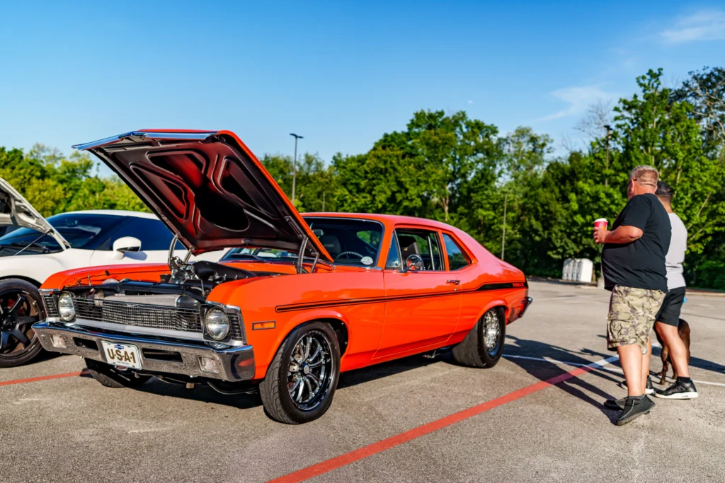 Orange Chevrolet Nova with hood open, parked in lot near two men standing and trees in background under clear sky