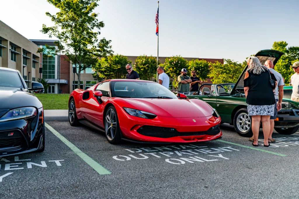 Red Ferrari 296 GTB parked next to a black Toyota GR Supra at an outdoor car event with people around and a green vintage
