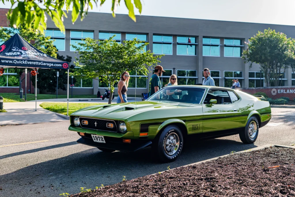 Green 1972 Ford Mustang Mach 1 parked outdoors with people around and a building in the background