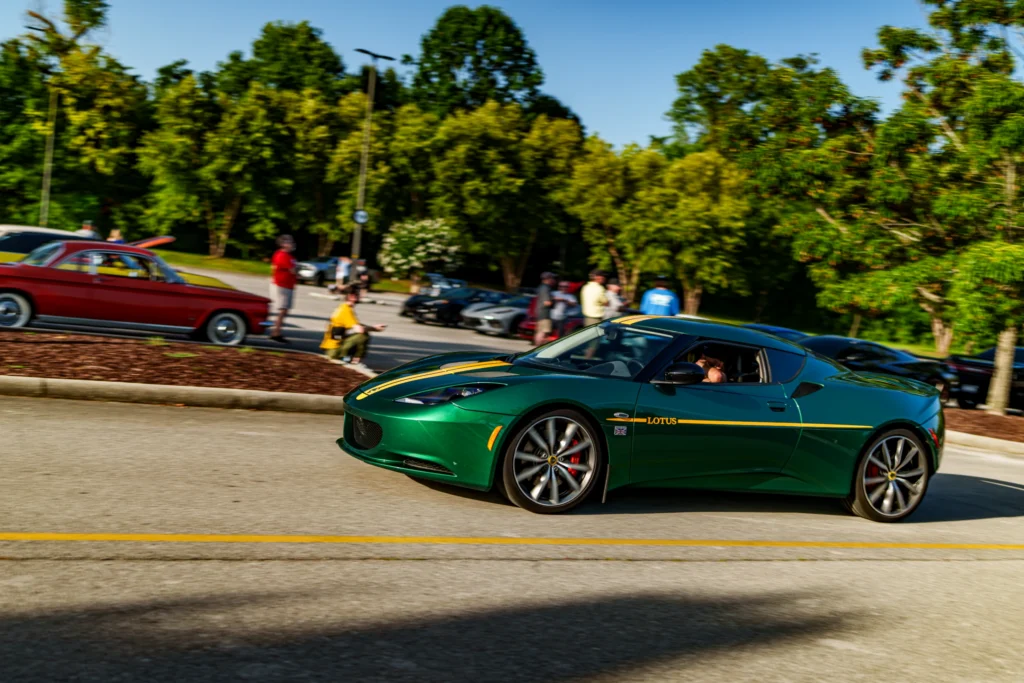 Green Lotus Evora S with yellow stripes driving on a road near parked cars and people in a parking lot with trees.