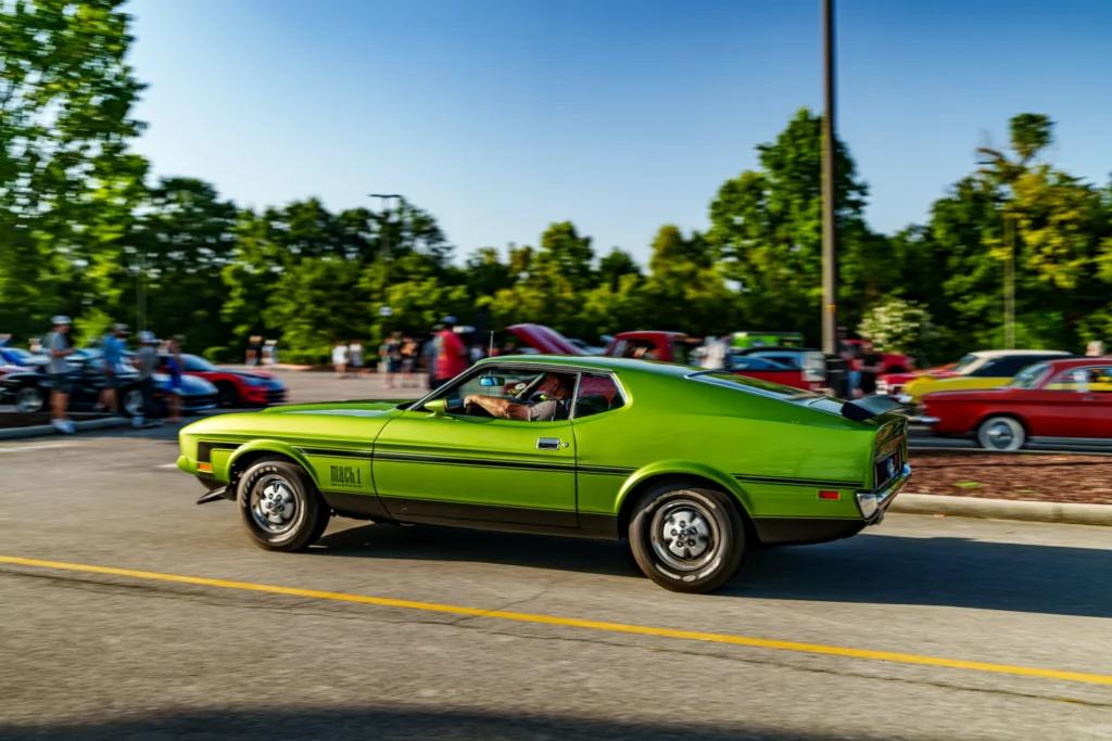 Green Ford Mustang Mach 1 driving on a road with people and parked cars in the background